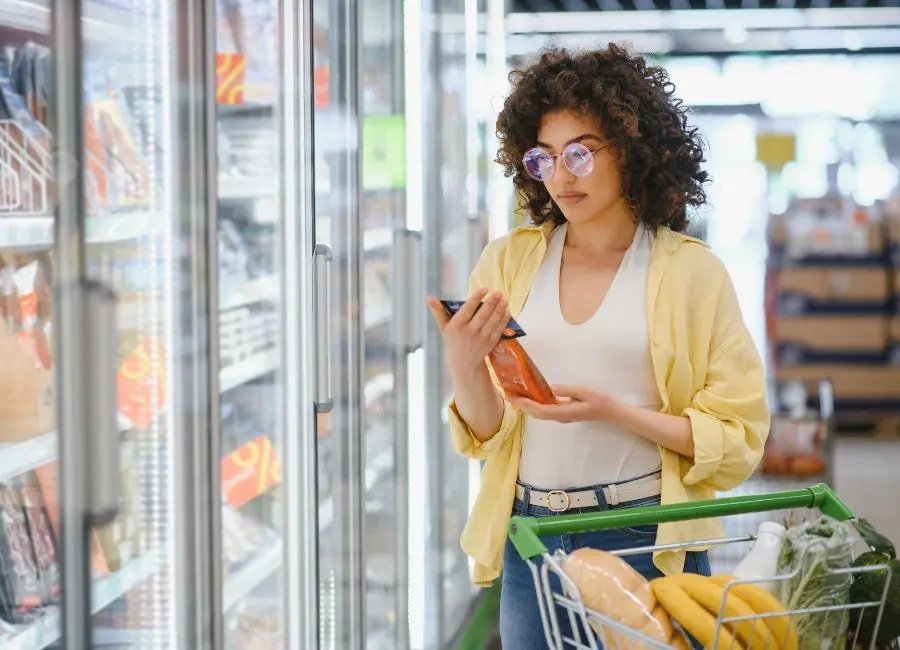 Young mixed race woman standing in grocery freezer aisle reviewing food label for nutrient content claims