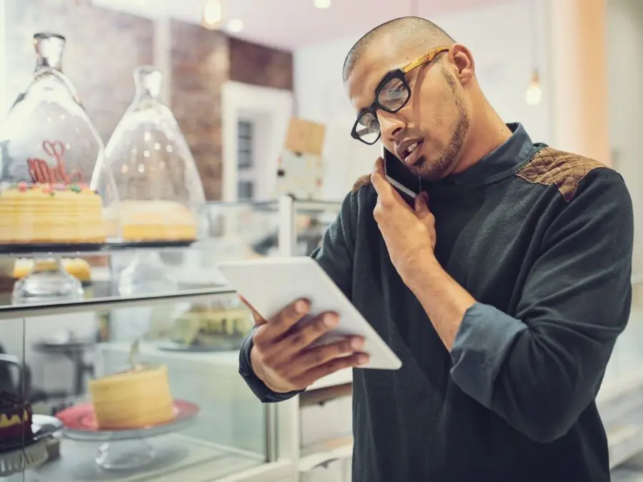 Cafe owner holding a tablet and talking on the phone beside a bakery display case with cakes, reviewing supplier orders in a modern coffee shop.