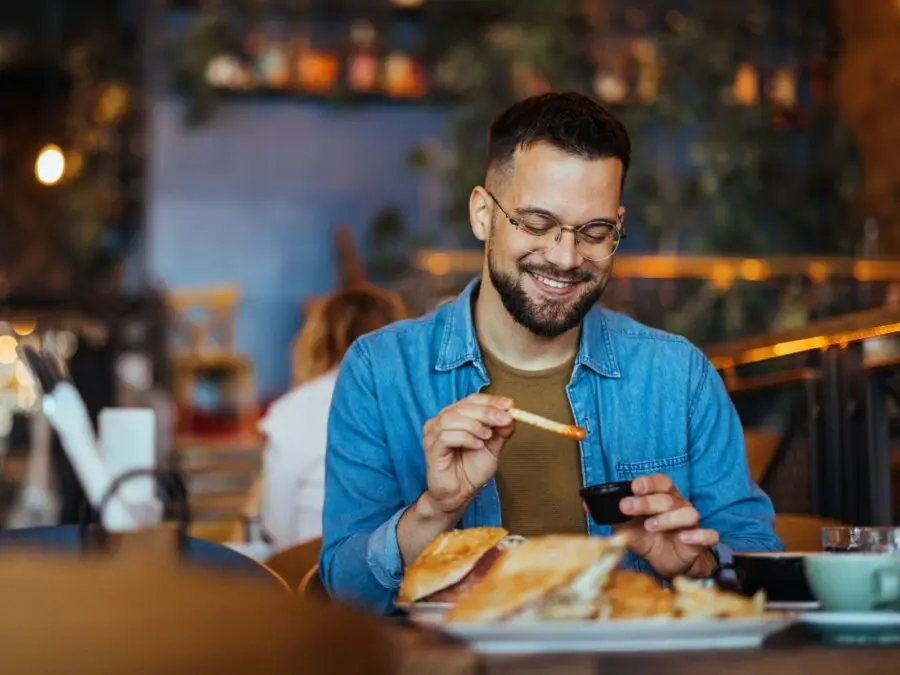 Smiling man dipping French fries into sauce while seated at a restaurant table, with burgers and a warm, softly lit dining background.