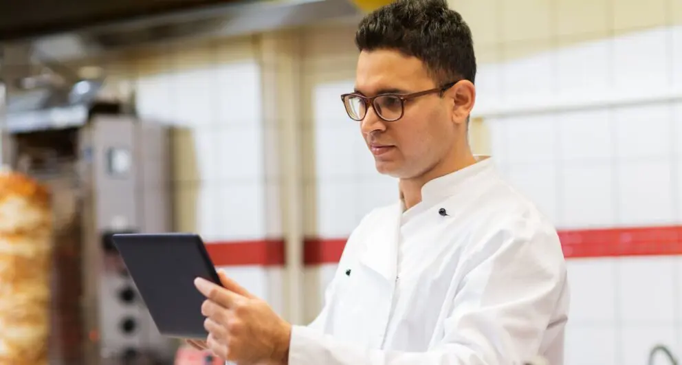 Chef using tablet in commercial kitchen to manage restaurant operations with digital software and smart food service technology.