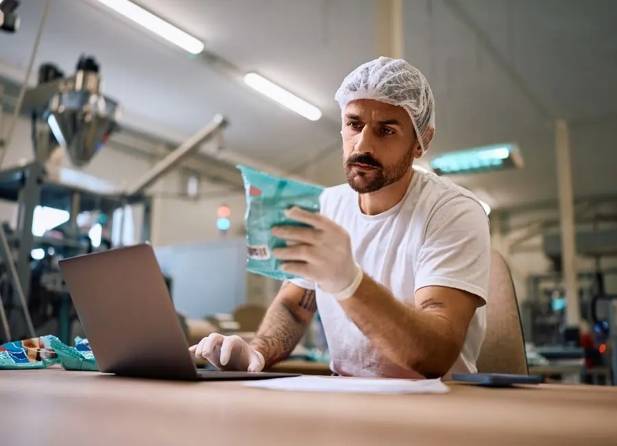 Food product manager in a hairnet and gloves inspecting packaged goods while working on a laptop in a production facility.