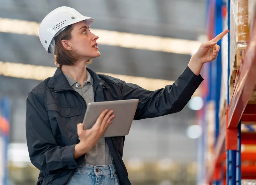Warehouse worker in a hard hat uses a tablet while inspecting packaged food on storage racks.