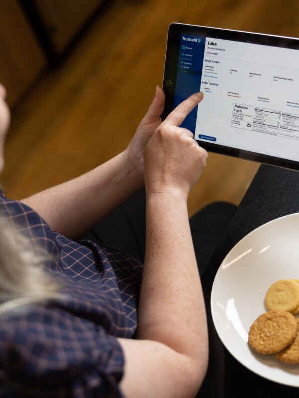 Woman using a tablet with a "Trustwell" label creation interface. A plate of cookies sits nearby.