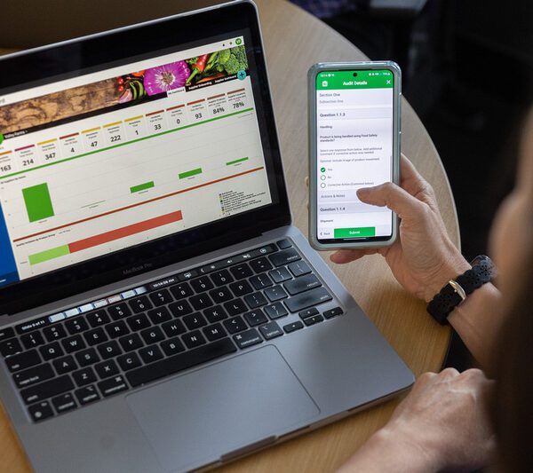 Person uses a phone to input data, while a FoodLogiQ by Trustwell dashboard is displayed on a laptop
