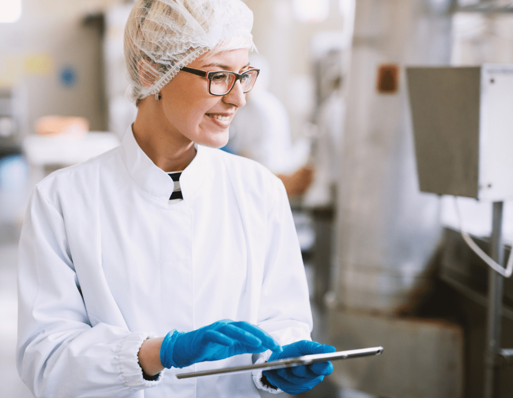woman in glasses, a labcoat, gloves, and a hairnet smiling as she holds a clipboard in a sterile manufacturing setting