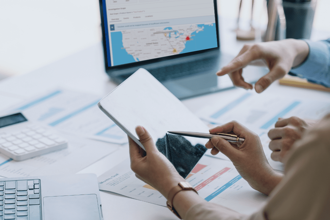Two professionals reviewing data on a tablet with supply chain maps and reports displayed on laptop and desk.