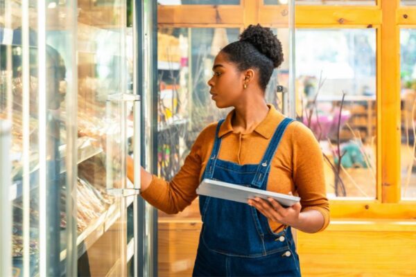 Woman in denim overalls using tablet to inspect frozen food in grocery store freezer