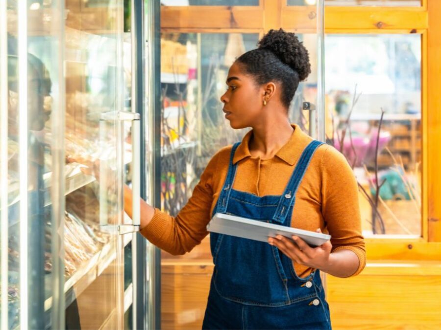 Woman in denim overalls using tablet to inspect frozen food in grocery store freezer