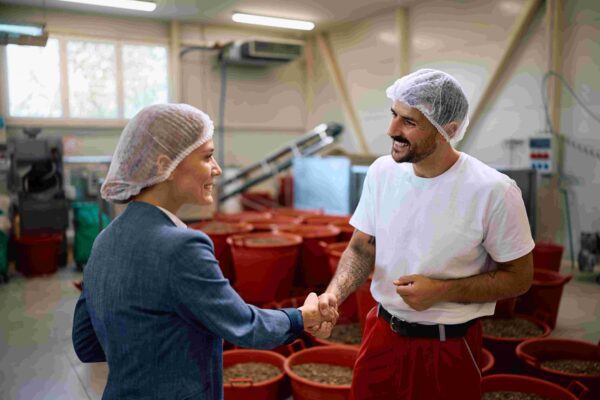 Two workers wearing hairnets shake hands in a food processing facility with red bins, showing partnership and quality control teamwork.