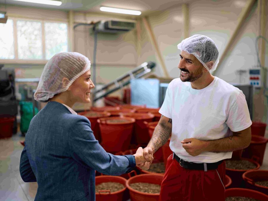 Two workers wearing hairnets shake hands in a food processing facility with red bins, showing partnership and quality control teamwork.