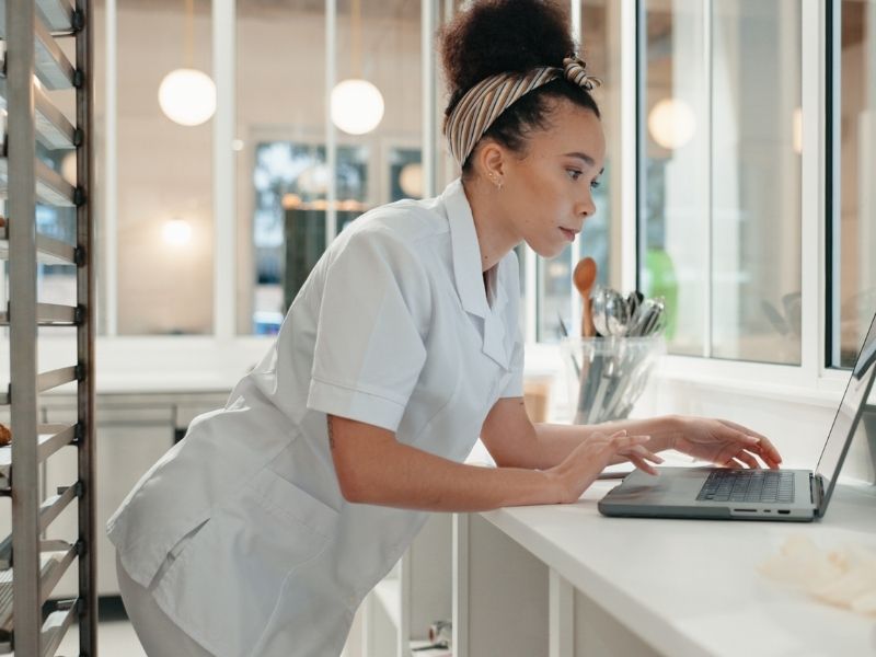 Focused young female baker in a white uniform uses a laptop in a modern, well-lit kitchen with utensils and baking trays visible in the background.