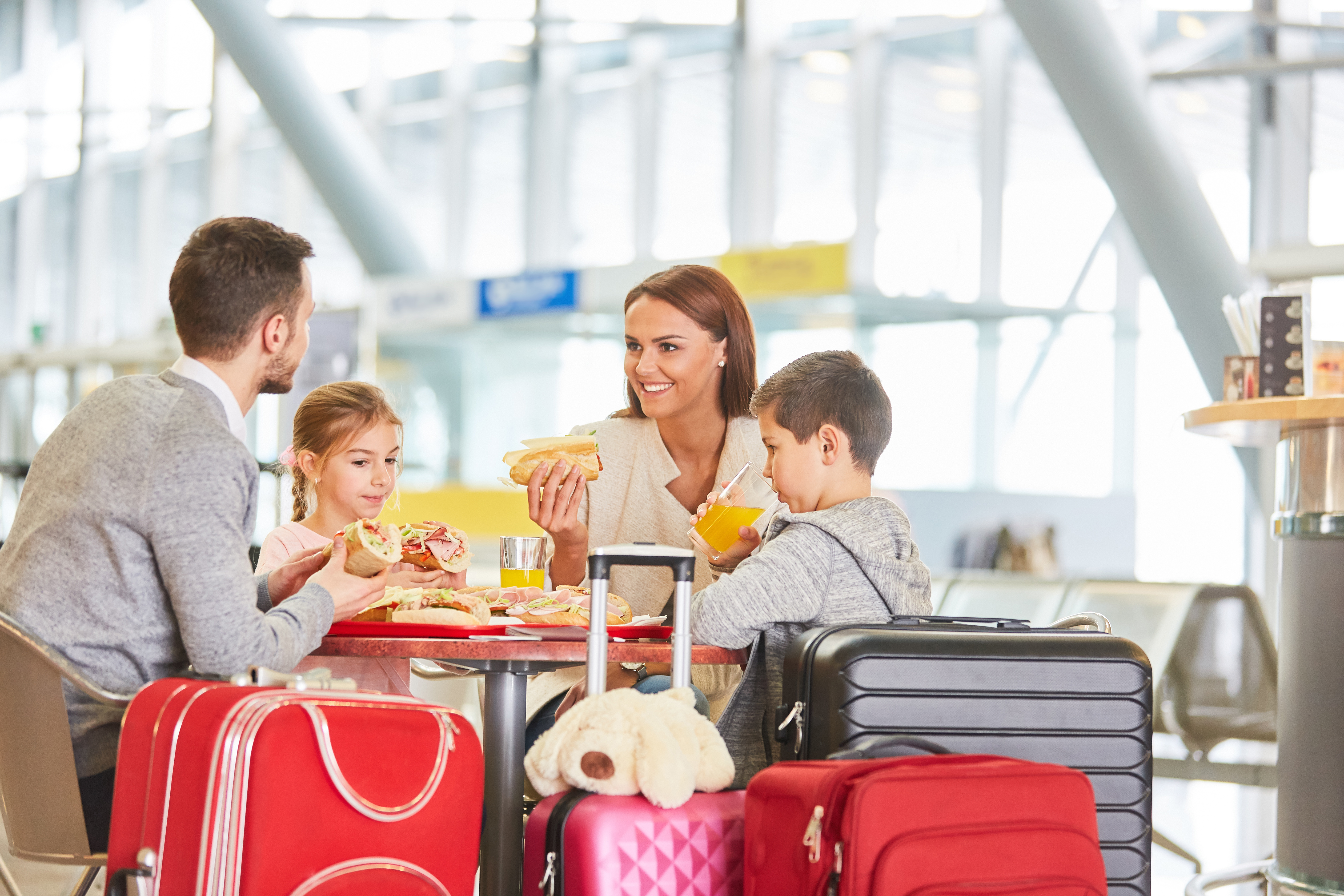 Family with two children eats sandwiches and drinks juice beside colorful luggage in a bright airport terminal.