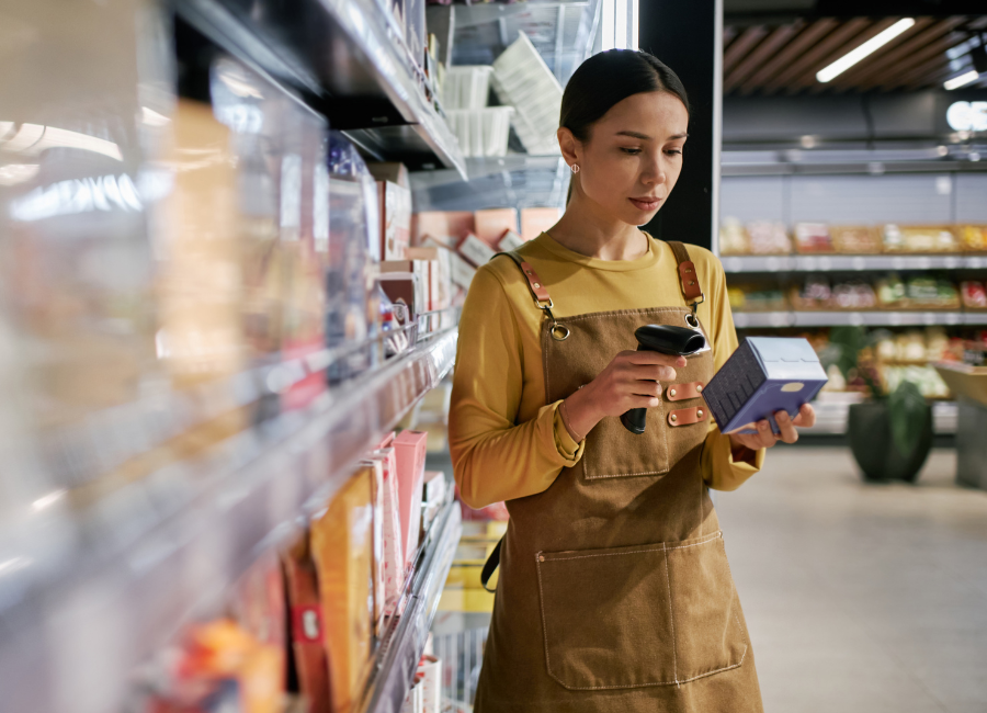 Grocery store worker uses a handheld barcode scanner to check traceability information on a packaged food product beside store shelves.